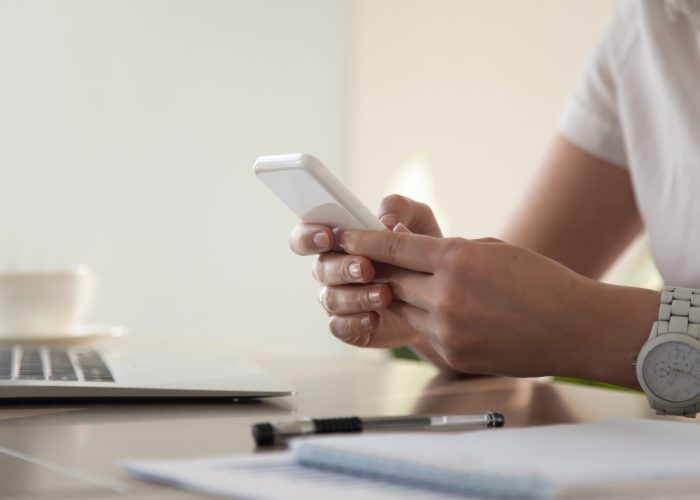 Close up image of modern cellphone in womans hand at desk with laptop. Office worker boring at workplace and playing games, writing in social networks. Businesswoman looking important contact on phone